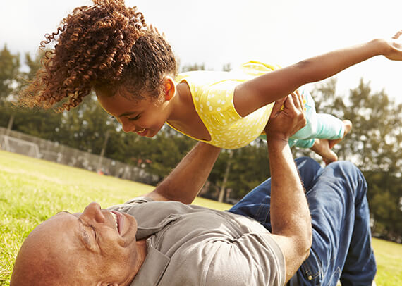 Man laying on back on the ground having fun holding granddaughter in an airplane pose.